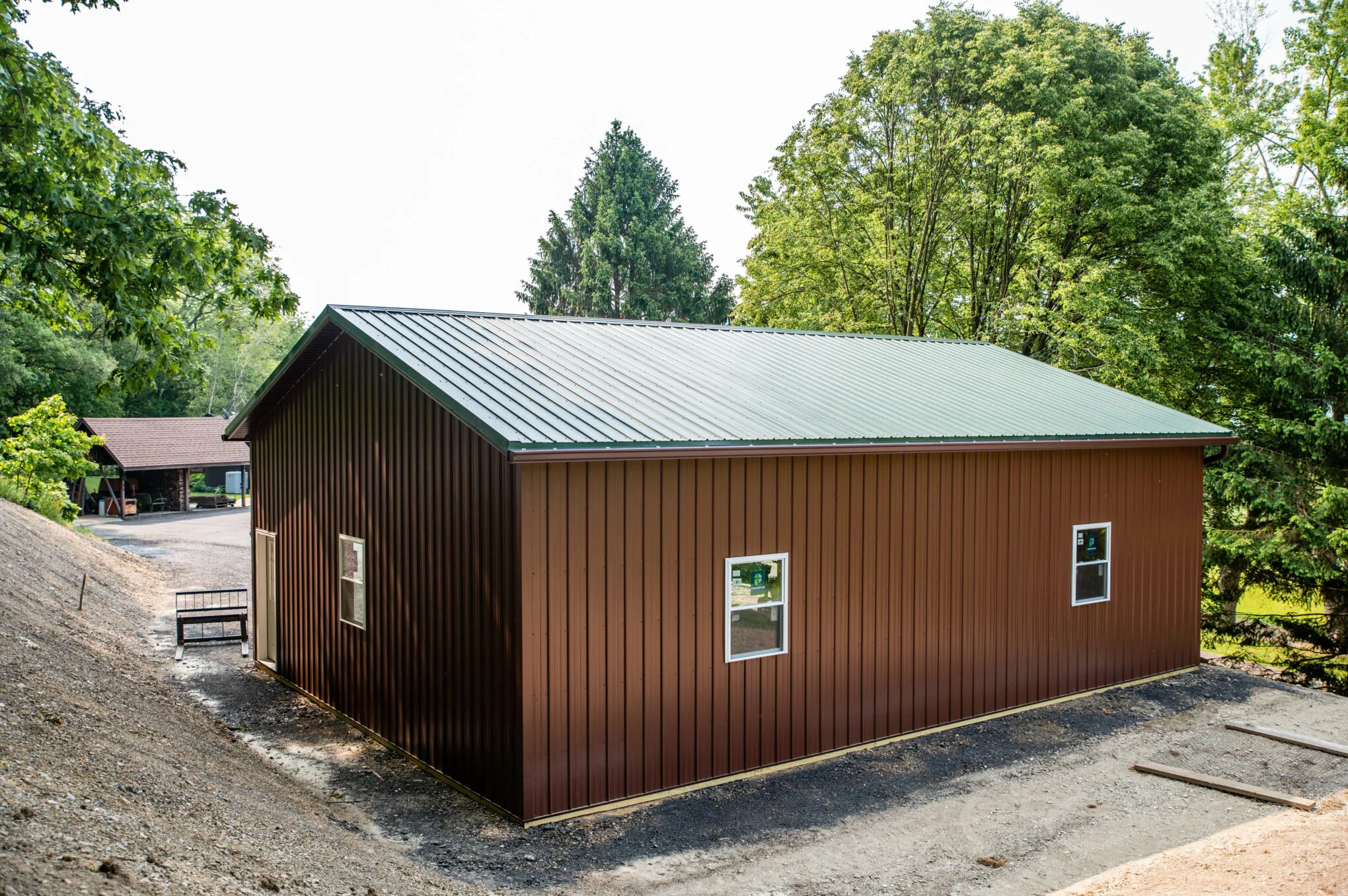 The gable end of a brown post-frame building with a green metal roof and white-framed windows, on a gravel pad